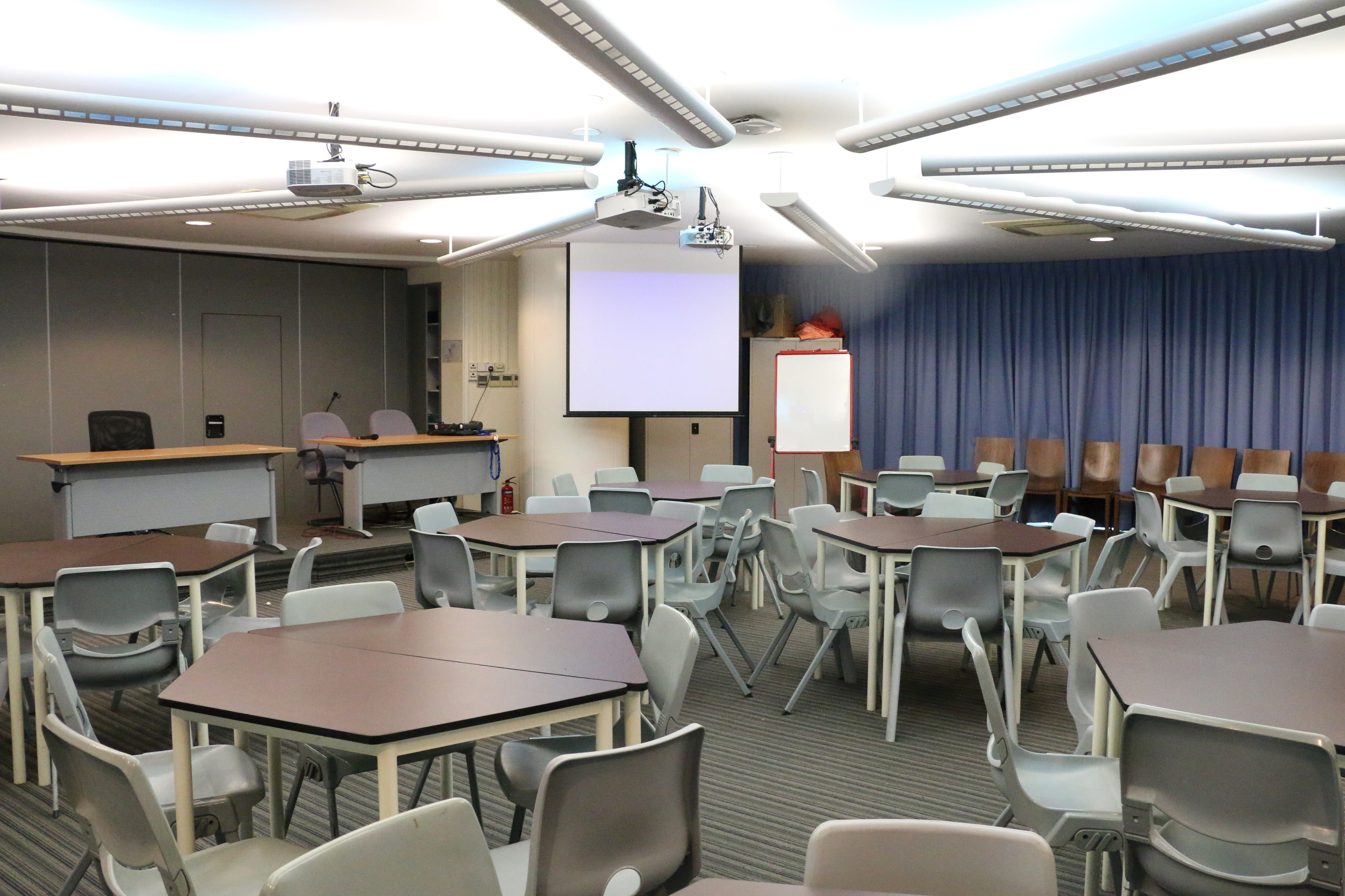 Empty classroom with round tables, gray chairs, two projection screens, and blue curtains along the walls.